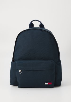 Navy blue fabric backpack with a rounded shape, front zip pocket, and a small logo patch. Features a top handle and adjustable straps.