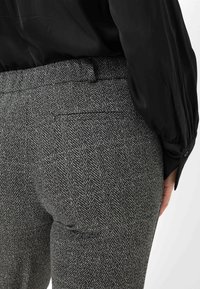 Grijze broeken met een visgraatmotief, gemaakt van textuurstof, met een achterzak en een aansluitende taille. Het model draagt een zwart topje.