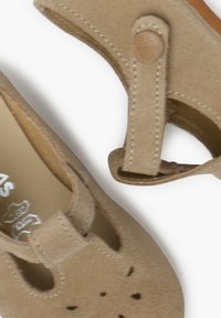 Beige ruskindssko med et rundt design, der har en rem med trykknaplukning, perforerede detaljer i tåområdet og en glat sål.
