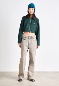 Chaqueta verde recortada con cierre de cremallera y bolsillo frontal, combinada con jeans de pierna ancha color beige. El modelo lleva un gorro azul y zapatos verdes.