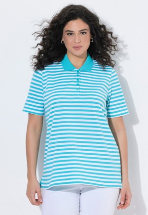 Kobieta o kręconych ciemnych włosach, ubrana w turkusowo-biały pasiasty polo oraz białe spodnie, stojąca na białym tle.