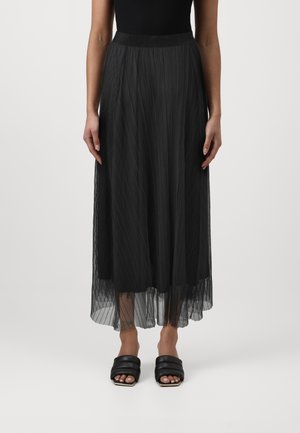 Femme portant une jupe noire plissée, taille haute, longueur cheville avec un voile transparent par-dessus, et des sandales à talons noires, ouvertes à bouts rembourrés.