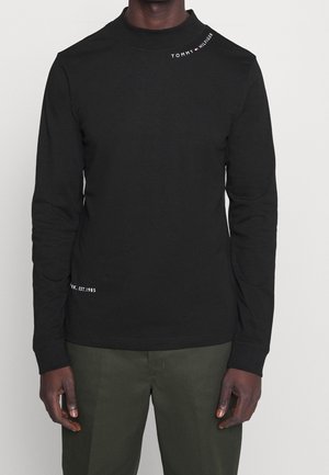 Mężczyzna ubrany w czarną koszulę z długim rękawem z logo Tommy Hilfiger przy kołnierzu oraz ciemnozielone spodnie, stojący na jednolitym tle.