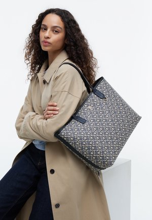 Vrouw met krullend haar, gekleed in een beige trenchcoat en donkere spijkerbroek, met een grote geprinte shoptas met zwarte handvatten over de schouder.