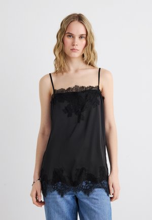 Mladá žena s vlnitými blond vlasmi má na sebe čierny top s čipkovaným lemom a modré džínsy, stojaca pred jednoduchým bielym pozadím.