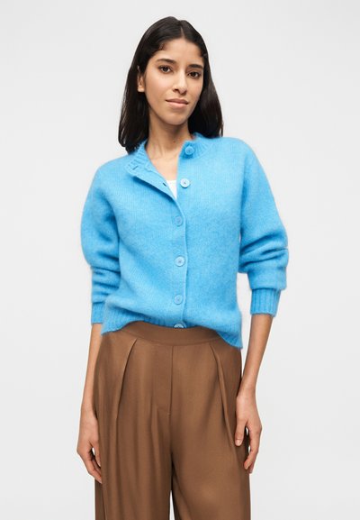 Γυναίκα με σκούρα μαλλιά που φοράει μπλε ζακέτα με κουμπιά πάνω από λευκό τοπ και καφέ, χαλαρά ψηλόμεσα παντελόνια, στέκεται μπροστά σε απλό φόντο.