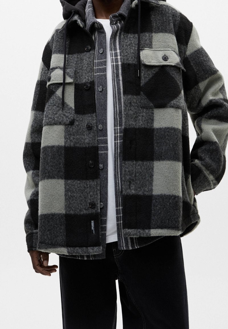 Homme portant des vestes à carreaux superposées noires et grises, dont une avec capuche et fermeture boutonnée, assorties à un pantalon noir.