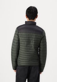 Polstret jakke i mørkegrøn med en sort øverste sektion, der har vandret quiltet design og høj krave. Båret med mørke jeans.