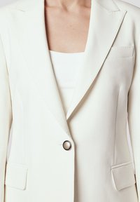 Biała marynarka o gładkiej teksturze, z klapami w kształcie notcha, dwoma przednimi kieszeniami i centralnym guzikiem o metalicznym wykończeniu. Idealna do eleganckiego ubioru.