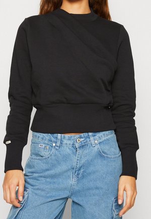 Vrouw draagt een aansluitend zwarte lange mouwen sweater en lichtblauwe denim cargobroek met hoge taille en zakken.