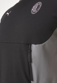 Camiseta atlética negra y gris con un alto cuello, mangas cortas, logo de Puma y escudo del AC Milan en el pecho. Tejido suave y texturizado.
