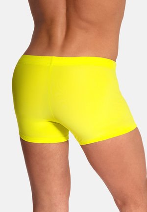 Henkilö, joka seisoo kirkkaan keltaisissa tiukasti istuvissa uima-alushousuissa ja jonka takana on vaalea tasainen tausta, nähtynä takaa.