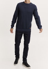 Pull en tricot bleu marine avec un col rond et des poignets côtelés, associé à un jean foncé et des baskets noires. Détail du tissu texturé visible.