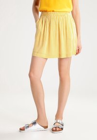 Jupe à rayures jaunes avec une ceinture élastique, offrant une coupe décontractée et des poches latérales, accompagnée de sandales argentées.