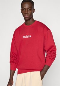 Piros hosszú ujjú pulóver, kerek nyakkivágással és a mellkas közepén fehér "adidas" logóval. Az anyag puhanak tűnik, bordázott mandzsettákkal.