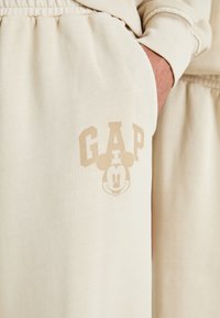 Beige jogger bikses, kas izgatavotas no mīksta materiāla, ar izcilu "GAP" logo un grafiku ar Mickey Mouse uz sānu kabatas.
