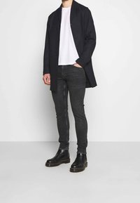 Svart lång kappa, vit t-shirt, mörka skinny jeans och svarta läderstövlar med kraftig sula. Slät textur, knappstängning framtill.