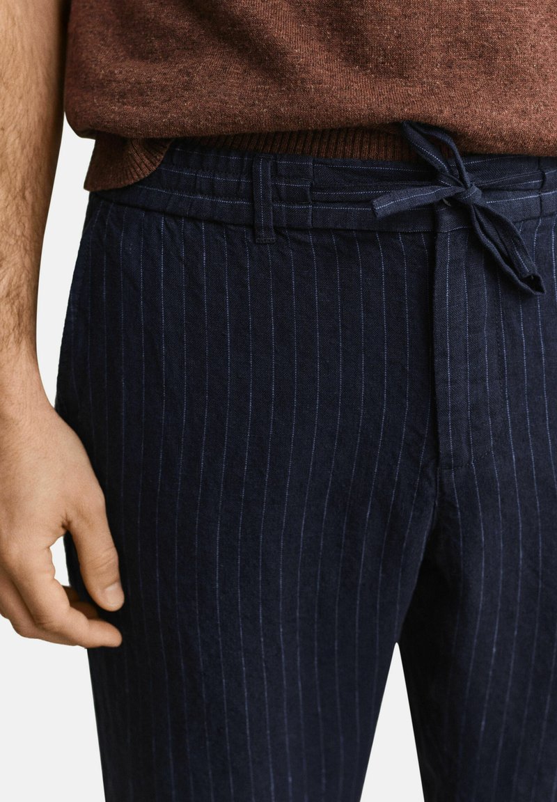 Mann trägt marineblaue Nadelstreifenhose mit gebundenem Bund und ein braunes Strickshirt, Hand entspannt an der Seite.