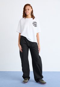 Weißes übergroßes T-Shirt mit schwarzem Logo, kombiniert mit lockeren schwarzen Hosen und olivgrünen Sneakers mit strukturierter Sohle.