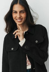 Tânără cu păr lung și închis la culoare zâmbește, poartă o jachetă neagră cu nasturi peste o cămașă albă cu text roșu, mâna aproape de bărbie.