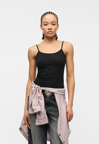 Νεαρή γυναίκα με πλεξούδες, φοράει μαύρο αμάνικο τοπ, ριγέ πουκάμισο δεμένο στη μέση, ζώνη με καρδιόσχημη αγκράφα και ξεβαμμένο μαύρο τζιν.