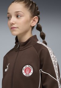 Chaqueta atlética marrón con acentos blancos, cuello alto, parche de logo y cinta estampada en las mangas. Cierre con cremallera en la parte delantera.