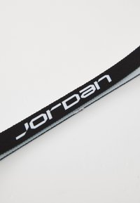 Correia de tecido preto com o logo branco "JORDAN" impresso em destaque. A correia apresenta uma textura suave e um leve brilho, afinando nas extremidades.