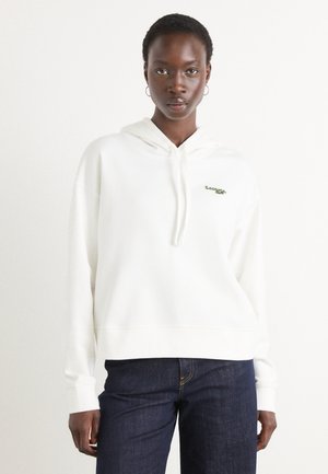 Hvit hettegenser med ribbestrikkede ermer og kant, med en grønn Lacoste-logo på brystet. Kombinert med mørkeblå denimjeans.