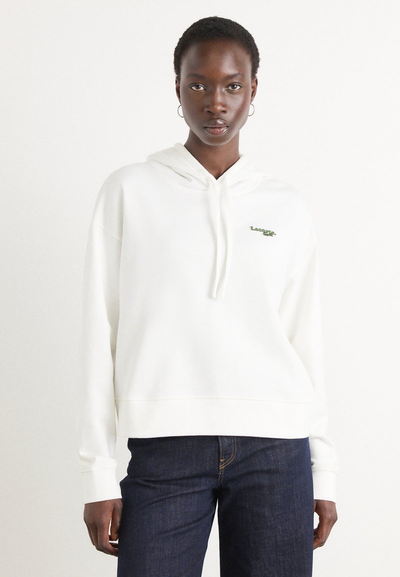 Vit hoodie med ribbade mudd och nederkant, prydd med en grön Lacoste-logga på bröstet. Matchas med mörkblå denimjeans.