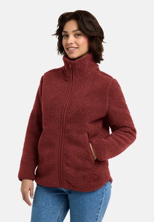 Woman wearing a zip-up, textured dark red fleece jacket with high collar and blue jeans, smiling with one hand in pocket.