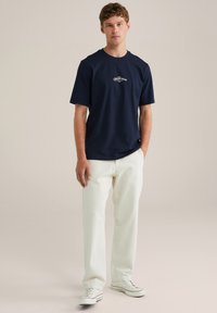 Marineblaues Baumwoll-T-Shirt mit kurzen Ärmeln und Rundhalsausschnitt; mit einem weißen Drucktext ausgestattet. Kombiniert mit cremefarbenen Hosen und Turnschuhen.