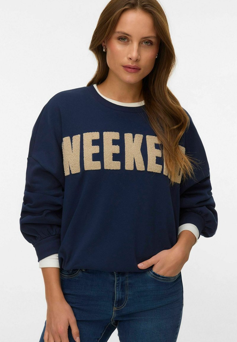 Femme aux cheveux longs bruns portant un sweat-shirt bleu marine avec le texte beige "WEEKEND", un jean bleu et des poignets blancs superposés, posant avec une main dans la poche.