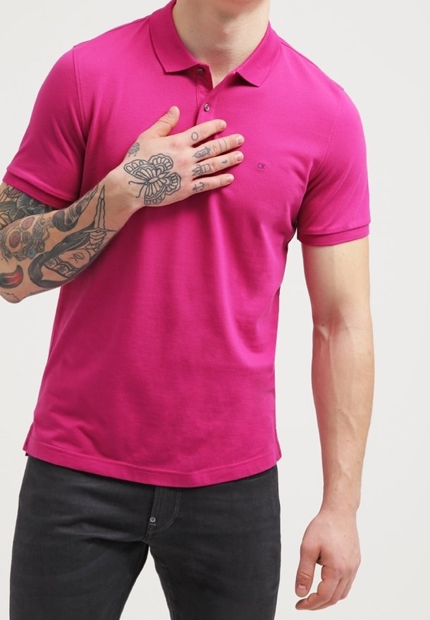 Homme portant un polo rose vif avec des tatouages sur le bras gauche et la main posée sur la poitrine, pantalon noir visible à partir de la taille.