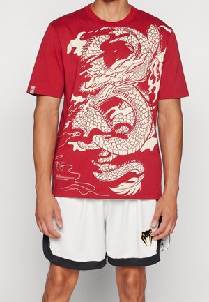 Homme portant un t-shirt rouge avec un grand motif dragon blanc et un short blanc avec une bordure noire et un logo serpent doré.