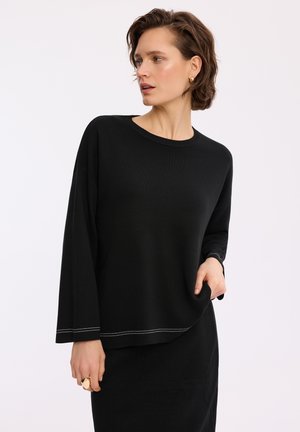 Kobieta z krótkimi włosami, ubrana w czarną bluzkę z długim rękawem z białym przeszyciem i pasującą czarną spódnicę, pozuje z jedną ręką w kieszeni.