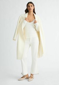 Top bianco a maniche lunghe con scollatura a girocollo, abbinato a pantaloni a campana coordinati e un cappotto color crema. Calzature con cinturini in evidenza.