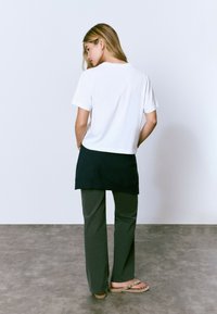 Camiseta blanca de manga corta superpuesta sobre una blusa negra, combinada con pantalones anchos de color verde oscuro y chanclas color beige, todo en un fondo neutro.
