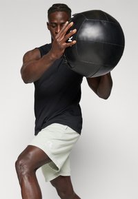 Black sleeveless top and light green shorts worn by a muscular person holding a textured black medicine ball, with a logo on the shorts.