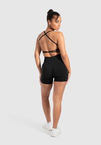 Zwarte sportbeha met kruisbanden op de rug, gecombineerd met hoge zwarte shorts. Model staat voor een neutrale achtergrond en draagt atletische witte sneakers.