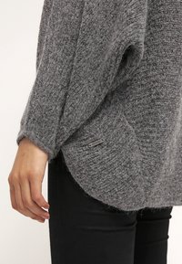 Szary dzianinowy sweter o fakturowanej tkaninie, w luźnym kroju, z logo na boku. Długie rękawy i zaokrąglony dół.