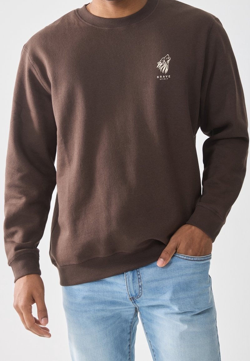 Homme portant un sweat-shirt marron avec un logo de loup et le texte « Brave Spirit », assorti à un jean bleu clair, main dans la poche.