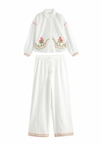 Valkoinen napitettava paita, jossa on vaaleanpunainen kukka-ommeltu kuvio, ja siihen sopivat valkoiset leveälahkeiset housut, joissa on kirjailutut hihansuut.