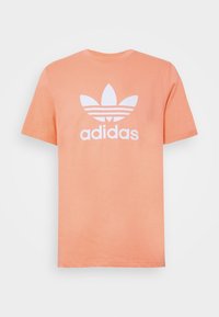 Persikofärgad bomullst-shirt med vit Adidas trefoil-logotyp och text. Kortärmad, rund halsringning, slät textur och regular fit.