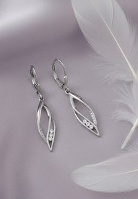 Silvriga örhängen med bladdesign, som har två cirkulära detaljer och dekorationer, mot en mjuk lila bakgrund.