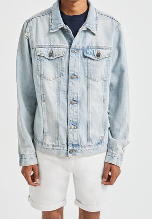 Mann trägt hellblaue Jeansjacke mit Knopfverschluss und weiße hochgekrempelte Shorts, steht vor einfarbigem Hintergrund.