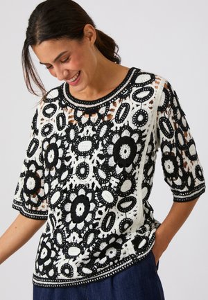 Donna sorridente verso il basso che indossa una maglia all'uncinetto bianca e nera con motivi floreali e maniche a tre quarti, abbinata a pantaloni scuri.