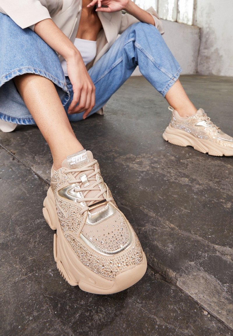 Personne assise par terre portant un jean bleu clair et des baskets épaisses beige décorées de strass et d'accents métalliques.