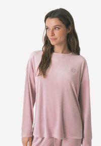 Roze langekleurige top gemaakt van zachte, geribbelde stof. Heeft een ronde hals en een klein zon-embleem op de linkerkant van de borst. Losse pasvorm.