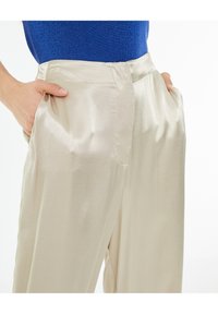 Pantaloni in satin color crema con una texture liscia, dotati di tasche laterali e una vestibilità comoda. Indossati con un top blu scintillante.