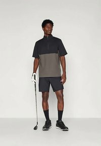 Πόλο κοντομάνικο με φερμουάρ σε σχέδιο χρωματικής μπλοκ σε μαύρο και γκρι, συνδυασμένο με σκούρες βερμούδες και μαύρα αθλητικά παπούτσια, κρατώντας ένα μπαστούνι του γκολφ.